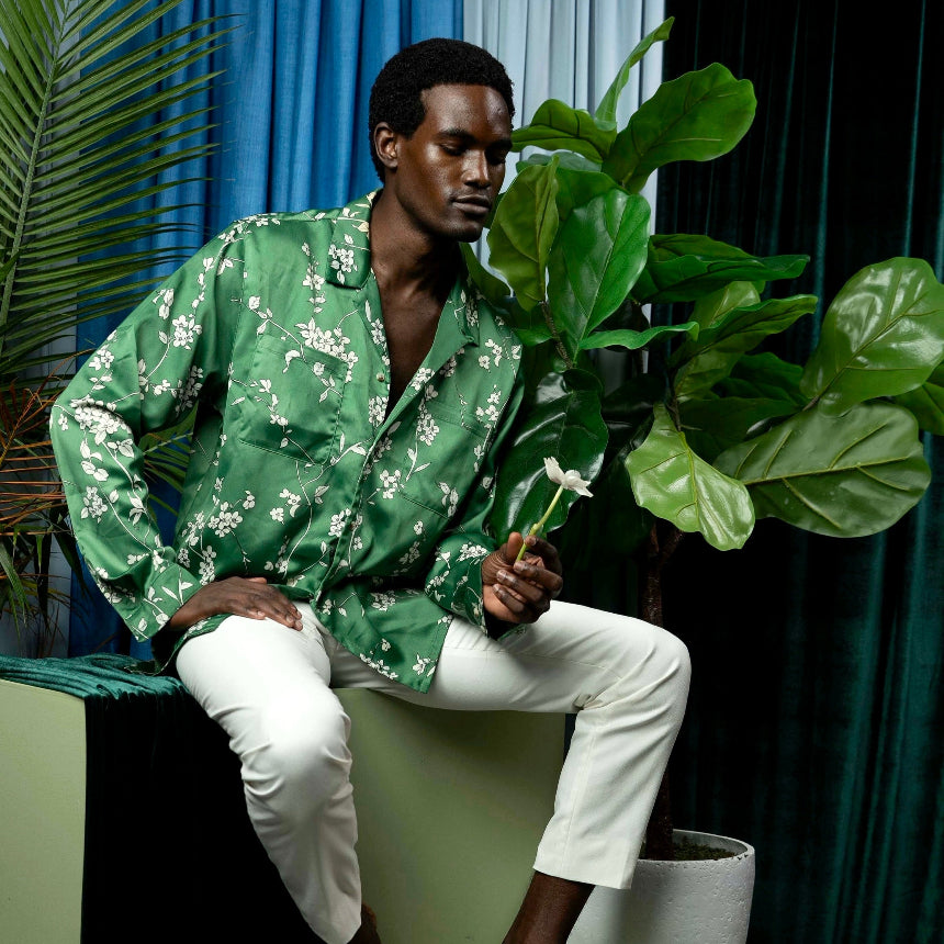 Man in a green patterned shirt and white pants sitting on a green box with plants around.